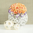Imagine a softly sunlit kitchen table in Mill Hill Village where a modern woven vase cradles a compact, dome-shaped bouquet that feels like a gentle sunrise. The arrangement is built around roughly two to three dozen peach and apricot roses, their petals layered and warm, exhaling a faint sweet fragrance that seems almost powdered into the air. These roses are arranged to show variety - fully open cups mingled with tight buds - producing a plush, rounded silhouette. Nestled along the lower curve, white Lisianthus bloom with creamy centres and edges painted in vivid violet; these ruffled, painterly blooms add a refined, slightly whimsical edge and lift the composition with delicate, translucent texture. Wisps of baby's breath (Gypsophila) are scattered throughout, lending a classic, airy veil that softens the overall density and creates tiny pockets of light. The flowers are set in a contemporary, white open-lattice basket that reads like woven reed or painted rattan - the bright white of the vessel contrasts softly with the warm rose tones and mirrors the pale, textured backdrop. Beside the base, two handwoven wooden spheres, one larger and one smaller, provide an organic accent and echo the basket's negative space. The result is romantic and understated: a centrepiece suited to anniversaries, welcoming housewarmings near Mill Hill Broadway, or a quiet celebration with close friends. As a florist who knows local tastes, I see it as a celebration of gentle sophistication and everyday joy.