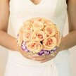 A softly lit, chest-level close-up captures a bride in a classic ivory gown cradling a round wedding bouquet, its colours glowing gently against the pale fabric. The lace bodice of her dress is delicately patterned, with a modest V-neckline and fine stitching that flows into smooth, layered folds at the waist, creating a timeless silhouette often seen in Mill Hill church weddings. Her hands, neatly manicured with pale, neutral polish and a faint shimmer on one thumb, rest tenderly around the stems, suggesting a moment of quiet anticipation before walking down the aisle. At the heart of the image is the bouquet itself, a compact dome of roses in soft peach, apricot and blush tones, their petals spiralling in tight, velvety whorls that speak of romance and warmth. Each bloom is nestled closely to the next, forming a plush, almost cloud-like surface of colour, with hints of coral pink deep in the centres. Beneath this rose canopy, a neat collar of ruffled purple blooms edged in creamy white, likely lisianthus, circles the base, adding depth and a gentle, unexpected contrast reminiscent of evening skies over Mill Hill Park. Around the lower edge, tiny sprays of white baby's breath peek through, airy and delicate, softening the transition between roses and skirt. The overall mood is serene and elegant, like a quiet pause outside a Mill Hill registry office or a hidden garden venue, and the bouquet feels freshly gathered and expertly arranged, ready to be carried into a day of celebration, photographs, and lifelong memories.