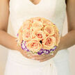 There is a gentle, poetic hush to this bridal bouquet, a look that reads like a summer memory gathered into a single, thoughtful bundle. At its heart sit richly formed apricot roses-creamy on the outer petals and warmer toward the centre-clustered symmetrically so each bloom supports the next, producing an organized, sunlit radiance. Surrounding the roses is a ring of dainty lisianthus, pale with violet tips and white throats, their delicate ruffled edges providing a cool, twilight contrast that frames the apricot core like a soft horizon. Interspersed among the main flowers is a fine lattice of tiny white baby's breath and slender green sprigs, lending an airy, weightless quality that suggests the bouquet might float on a warm breeze. The petals' surfaces show subtle veins and a soft, satiny sheen under gentle daylight, while shadows fall with a hushed softness that accentuates the arrangement's layered depth. This design is the kind I often prepare for weddings around Mill Hill Broadway and Mill Hill Village, where brides ask for something refined, timeless, and comforting. On the nose it reads as tender and fresh, a quiet mix of sweet rose perfume with green, leafy notes. Delivered with care from Flowers Mill Hill, the bouquet is made to sit lightly in the bride's hands, to complement lace and silk without shouting, and to become part of a day built from careful, loving details and small, resonant moments.