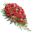 This image shows a grand, elongated funeral spray in deep red tones, displayed on a spotless white background that sets off its shape and colours with clarity. The arrangement sweeps diagonally from the upper left to the lower right, wider and more rounded at the top and narrowing into a refined, pointed tip, echoing the familiar outline of a casket spray. At its centre, velvety deep red roses are fully open, their petals overlapping in soft, sculpted spirals, each flower carrying an air of solemn grace. Between them, smaller red spray carnations and carnations in a slightly brighter shade contribute fine, fringed petals and layers of texture, ensuring there are no gaps and that the spray feels lush and complete. Supporting this mass of flowers is a thick cradle of greenery, with broad, dark green leaves forming a robust foundation that extends beyond the blooms to define the outline. Dotted among the deeper greens are rounded stems of silvery-green eucalyptus, whose cooler hue and gently matte finish bring a quiet, calming balance to the richness of the reds. At the lower right where the spray narrows, strands of light green trailing foliage arc outward and downward, adding a sense of natural movement and drawing the eye along the full length of the tribute. The lighting is soft and even, highlighting the subtle sheen of the petals and the varied textures of the leaves. This is the kind of traditional funeral display often seen in services around Mill Hill, a thoughtful floral expression of remembrance, sympathy, and lasting respect crafted to accompany a final goodbye.