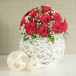At the heart of this scene is a dome-shaped bouquet of approximately a dozen red roses, each bloom unfurling into a lush, velvety spiral that immediately suggests romance and celebration. Their colour is a deep, almost ruby crimson, rich but not overpowering, creating a smooth expanse of petals that feels both classic and modern. Intermixed with the roses, slender stems of pure white gypsophila weave their way through, the minute blossoms forming a whisper-light veil that softens the bold palette and adds a lace-like texture. Around the base of the dome, small touches of glossy dark green foliage emerge, grounding the composition with a hint of natural woodland tones. The arrangement is cradled in a spherical vase constructed from white woven strands, its open, crisscross pattern resembling contemporary basketry. The spaces between the weaving allow light to slip through and create subtle patterns on the surface below, enhancing the piece's sculptural quality. Two decorative balls, made from similar pale, natural fibres and differing slightly in size, sit on the crisp white tabletop beside the vase, echoing the sphere motif and tying the décor together. The backdrop is a softly textured wall in a muted beige or light grey, simple and unobtrusive, deliberately chosen to let the bouquet's rich colours shine. Soft, diffused lighting washes evenly over the entire arrangement, revealing every contour of petal and every strand of woven material without harsh glare. The composition feels perfectly suited to a refined wedding breakfast in Mill Hill, perhaps near Mill Hill Park, or as a special gift delivered across NW7, where a bouquet like this from Flowers Mill Hill would speak quietly but confidently of love, commitment, and thoughtful design.