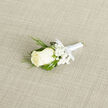 Viewed up close, this boutonniere presents a study in subtle contrasts and refined texture. An immaculate creamy-white rosebud commands the centre, its velvety petals arranged into a soft, compact spiral. Around it, delicate clusters of baby's breath provide an ethereal, almost cloudlike softness, their tiny white pinpricks of blossom suggesting lightness and breath. Complementing these soft elements are narrow, feathery sprigs of dark green foliage-evocative of cedar or mature evergreen-that add depth and a whisper of forest coolness, while fuller deep-green leaves behind them give the piece structure and visual weight. A slim white satin ribbon binds the stems, tied into a neat bow whose satin sheen plays gently with the light on the woven beige fabric beneath; the fabric's subtle texture frames the boutonniere as if on a small stage, emphasising each petal and frond. The overall scent seems quietly floral-clean, faint, and uplifting-suitable for the intimacy of a wedding aisle or the hush of a prom reception. As an experienced florist working in and around Mill Hill Village and The Ridgeway, I design boutonnieres like this to complement classic suits and softer colour palettes, often matching flowers used in bridal bouquets from nearby ceremonies. It reads as timeless and thoughtful, an accessory that carries both the care of the maker and the gentle promise of the day it commemorates.