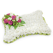 A plush, rectangular memorial pillow completely cloaked in hundreds of tiny, pristine white chrysanthemums forms the soft, fabric-like surface of this sympathy tribute. The closely clustered blooms create a smooth, cushiony texture that suggests quiet rest; from a short distance the pillow reads as an unbroken field of white, while up close individual petals and the faint, fresh scent of chrysanthemum and freesia are discernible. Set delicately on the upper-left corner is a focused spray of rich fuchsia and deep magenta roses, their velvety petals unfurling in concentric layers that draw the eye and offer a warm counterpoint to the tranquil white. Interspersed among those roses are small, pure white freesias or buds, their slender throats and delicate petals adding lightness and a hint of soft perfume. Lush greenery - slender fern fronds and glossy leafy sprigs - fans beneath and around the accent cluster, giving it a natural frame and depth against the snowy pillow. Around the pillow's edge a pleated ribbon of pale green satin peeks out like a subtle halo, its gentle sheen and looped finish neatly defining the rectangular form and suggesting renewal. The arrangement sits against a clean white backdrop with a soft shadow beneath, emphasising three-dimensional shape and the careful craftsmanship of the florist. Intended as a funeral or graveside tribute, this classic pillow speaks of peace, reverence and lasting remembrance - a tactile, visual comfort for those gathering to honour a life in and around Mill Hill, where local chapels and memorial gardens often receive such quiet, dignified tributes.