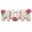 A tender, standing memorial piece that spells the word MUM in three large, connected block letters, photographed against a plain white backdrop. Each letter is lovingly constructed from dense clusters of pristine white chrysanthemums, their small, feathery heads packed tightly to create a soft, pillowy surface that reads as gentle purity and quiet dignity. The outer edges of every letter are softly finished with a ruffled pale pink ribbon or fabric trim, which catches the light and lends a warm, motherly halo to the otherwise white composition. A slim, dark green support frame peeks through between the letters, giving a stable silhouette and a subtle contrast to the whiteness. At three carefully chosen points-upper left of the first M, the inner curve of the U, and the upper right of the final M-sit compact posies of vivid fuchsia-pink roses, glossy green fern-like foliage, and clusters of reddish-orange hypericum berries; these vibrant accents break the white plane with sympathetic colour and textured interest. The overall scent is cupped and soft: the cool, green undertone of chrysanthemum mat, the velvet perfume of roses, and the faint tang of berried foliage. The composition reads as a respectful, personal farewell - ideal for a funeral, wake, or graveside dedication - its materials and finish reflecting careful florist craft and an intention to comfort. As if arranged by a thoughtful local florist in Mill Hill, the piece combines classic sympathy florals with a small modern twist of colour, offering a composed and heartfelt tribute to a cherished mother, ready to stand at a service or beside a graveside on a quiet morning.
