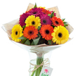 A festive and vibrant bouquet of Gerbera daisies catches the eye, each blossom radiating warmth and cheer reminiscent of Mill Hill's welcoming avenues. The arrangement dazzles with its blend of sunny yellows, luminous oranges, and bold magentas, their flat petals elegantly fanned out around dramatic dark centers. Among the riot of blooms, lush green leaves weave through, adding organic structure inspired by the peaceful greenery near Mill Hill East. The stems are grouped with precision and showcased in a clear glass vase, its water crystal clear and life-giving. Wrapped in a rustic, lightly textured brown kraft paper and finished with an airy cellophane cone, the bouquet's packaging merges countryside charm with practical elegance. A natural twine tie encircles the stems, from which dangles a neat white tag featuring a delicate red rose-an emblem of thoughtful giving. Illuminated under natural studio lighting, each color pops vibrantly, creating a mood of celebration and heartfelt intent. Ideal for marking a milestone, expressing thanks, or brightening a loved one's day in NW7, this arrangement exemplifies the artistry and warmth at the heart of Mill Hill's vibrant community, turning any occasion into a memorable affair.