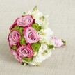 A vibrant and elegant bridal bouquet lies against a subtly textured light beige fabric, photographed from a slightly elevated angle so the rounded shape can be clearly read by the eye. At the centre, several fully opened roses in a soft lavender-pink gradually deepen toward a richer fuchsia along the petal edges, creating a romantic two-tone swirl that feels both modern and timeless. Between these lush blooms, small chartreuse green button chrysanthemums pop like tiny fireworks, their tight, domed heads adding crisp texture and a cool freshness that contrasts beautifully with the velvety rose petals. Delicate white flowers, likely lisianthus, are woven throughout the bouquet, their ruffled, layered petals and pale greenish-yellow centres bringing a cloud-soft lightness that softens the whole design, much like early spring skies over Mill Hill Park. A few small, closed green buds peek out here and there, suggesting just-cut, garden-fresh stems, as if they've only just arrived from a local grower on Mill Hill Broadway. The stems are neatly gathered and finished in a decorative handle, wrapped in smooth pink satin ribbon that catches the light with a gentle sheen. Over this, a fine strand of small, lustrous white pearls spirals down the handle, echoing heirloom jewellery and giving the bouquet an unmistakably bridal character. The composition feels perfect for a Mill Hill wedding ceremony, a civil service at a nearby venue, or as inspiration for sophisticated event flowers, with its harmony of pink, white and green offering a visual story of romance, celebration, and thoughtful floral craftsmanship.