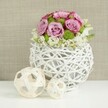 This image presents a stylish wedding-style table arrangement, as if set for a celebration in one of Mill Hill's elegant homes overlooking green, tree-lined streets. The focal point is a spherical white vase fashioned from intertwining, twig-like rattan strands. These strands loop and weave around each other, leaving gaps that create an airy lattice, giving the vessel a sculptural, modern feel. Rising from this openwork base is a lush, domed bouquet of flowers. Soft pink and lavender roses dominate, their petals unfolding in generous layers and edged with subtle deeper pink tones that add romance and dimension. Tucked between the roses, bright green button chrysanthemums stand out with their small, neatly rounded forms and finely textured petals, offering a crisp contrast to the roses' smooth curves. Gathered at the base of the dome, delicate white filler flowers cluster and dip slightly over the rim, creating a gentle halo and enhancing the sense of fullness. On the smooth, white tabletop to the left of the vase sit two decorative spheres made from pale wicker, one larger and one smaller, both featuring an open, crisscross pattern echoing the vase's weave and adding visual rhythm. The background is softly blurred in warm beige tones, reminiscent of natural linen, which keeps the focus on the arrangement while adding a subtle sense of depth and warmth. Soft, even light highlights the texture of the petals and the intricacy of the wicker, giving the composition a calm, refined mood. It's an image that captures the quiet luxury of fresh flowers prepared with care by a local Mill Hill florist for weddings, anniversaries, or intimate gatherings.
