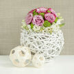 A stylish floral arrangement takes centre stage on a bright white, reflective tabletop, its surface catching soft glimmers of light as if from a nearby Mill Hill window. The focus is a compact, dome-shaped bouquet of pink roses and lime green button chrysanthemums, elegantly presented in a spherical vase made from interwoven, white-painted branches. This open, twig-like structure has the feel of a handcrafted sculpture, its airy design echoing the green, spacious character of Mill Hill's gardens and parks. The roses are luxuriously full, petals gently spiralling outward in shades of blush and lavender-pink, some with slightly deeper edges that add depth and romance, making the piece ideal for a wedding reception or engagement dinner. Nestled among them are round, button-sized chrysanthemums in a vivid chartreuse tone, their numerous tiny petals creating a textured contrast that brings freshness and a hint of playfulness. Encircling the base of the arrangement, a fringe of creamy-white flowers and unopened buds drapes softly over the rim, their delicate stems and faint yellow centres providing a cloud-like transition between the structured dome and its airy container. To the left, two decorative balls crafted from pale wooden strips rest on the table, their open lattice pattern subtly mirroring the woven vase and reinforcing the natural, artisanal theme. The background is a warm, taupe-toned wall with a fine, linen-style texture, blurred enough to keep the eye firmly on the flowers while adding cosiness and calm. The overall impression is of understated yet polished elegance-a perfect centerpiece for Mill Hill weddings, intimate celebrations, or tastefully styled homes where gentle colour and careful floristry are cherished.