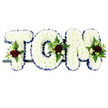 The photograph captures a classic name tribute for a funeral, with the word TOM crafted in three large floral letters that rest on a slender dark frame. Each capital letter is solidly formed from compact clusters of white chrysanthemums, their small, textured petals overlapping to create a soft, uniform surface reminiscent of fresh snow. This carpet of white stands out clearly against the plain white background, giving the tribute a serene, almost weightless quality that would look at home in a chapel or crematorium serving the Mill Hill community. Outlining each letter is a border of royal blue satin ribbon, neatly pleated and pinned so that the rich blue hue and gentle sheen frame the chrysanthemums like a tailored suit. At specific points on each letter, small, thoughtfully designed sprays of accent blooms bring depth and colour: deep, velvety red roses, smaller white or cream spray roses, and slender white flowers with a freesia-like form, all nestled among fresh green foliage. Fern-like fronds and glossy leaves extend slightly beyond the letters, softening the edges and adding a sense of natural movement, as if a breeze from nearby open spaces had passed through. The three letters are arranged horizontally, clearly intended as a sympathy tribute to sit alongside a coffin or be placed at the front of the service. The overall design carries a quiet dignity and tenderness, offering a personalised farewell for someone named Tom - a floral message of love, remembrance, and peace from those gathering in Mill Hill to say their last goodbyes.