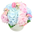 A beautifully composed hatbox bouquet in pastel tones stands out against a crisp white background, its round white container giving the design a clean, contemporary feel. The box itself is simple and elegant, with a faint ridge wrapped around its middle, while the flowers above form a soft, overflowing dome. At the centre, large pink peonies draw the eye, their plush, many-layered petals creating a sense of romance and gentle movement, as though each blossom is just about to expand further. Interlaced with the peonies are full heads of light blue hydrangeas, their countless small florets coming together into cloudlike clusters that cool the palette and evoke peaceful skies over Mill Hill's parks and gardens. Scattered throughout are dainty light pink spray roses in various stages of bloom, from tightly curled buds to small open flowers, adding intricate detail and a sense of unfolding stories. Two vivid pink calla lilies rise from within the pastel mound, one reaching towards the upper left, the other towards the right, their smooth, trumpet-shaped forms introducing a striking, modern contrast to the ruffled petals around them. A few rich green leaves peek from the base, offering depth and anchoring the composition. The arrangement is lush and generously proportioned, suggesting celebration and care. It feels perfectly suited for marking personal milestones in Mill Hill-perhaps a new baby welcomed in a nearby family home, a special birthday, or a refined sympathy gesture-bringing a touch of soft luxury and calm beauty to any local setting, from a smart townhouse off Mill Hill Broadway to a cosy apartment tucked away on a quiet side street.