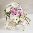 A romantic, basket-based floral centerpiece is styled as though ready to be placed at the heart of a wedding table in Mill Hill, captured against a soft, neutral fabric backdrop that gently fades into the distance. At its heart, several generously open pink roses display their intricate spiral of petals, evoking affection and refined beauty. These are surrounded by striking white Phalaenopsis orchids, whose wide, graceful petals and sunny yellow centers introduce an elegant, contemporary note, reminiscent of chic city weddings. Between them, white Lisianthus blooms with delicately ruffled petals, some edged in the softest lavender, create a tender, layered texture. Pale pink stock stems rise delicately and then slope downwards, softening the edge of the rounded white wicker basket and lending a subtle cascading effect. Airy clusters of baby's breath dot the arrangement like scattered pearls, giving the design a light, cloudlike quality. Fresh green foliage threads through the composition, with leaves that peek between blooms and occasionally trail outward, echoing the greenery of nearby Mill Hill gardens and tree-lined streets. The basket itself, tightly woven and painted bright white, rests firmly on a clean white surface that enhances the arrangement's pastel tones. To one side, two decorative spheres made from fine, entwined wooden strands sit side by side, their natural beige hues complementing the basket's rustic charm. The background, a calm beige textile with gentle texture, stays quietly in place so that the flowers and foliage remain the undisputed focus. The overall impression is of soft luxury and heartfelt celebration, ideal for showcasing wedding table flowers, engagement party decor, or refined event arrangements available from Flowers Mill Hill across the local area.