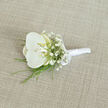An elegant white orchid boutonnière is carefully arranged on a pale beige, linen-textured fabric, the whole composition exuding the calm, polished atmosphere of a Mill Hill wedding morning. At the heart of the piece, a single orchid bloom opens generously, its broad, velvety petals a luminous white that almost glows against the neutral ground. Inside the flower's centre, a soft blush of cream and pale yellow adds depth and a whisper of warmth, drawing attention to its beautifully defined throat. Encircling this focal bloom, airy sprigs of white baby's breath create a halo of tiny blossoms, like a fine, floral mist that softens the line between the orchid and the space around it. Slender, fresh green blades of foliage, echoing the clean, linear greenery you might notice along the paths near Arrandene Open Space, extend lightly from beneath the flowers, introducing movement and a subtle sense of energy. The stems are expertly bound and wrapped in a smooth, glossy white satin ribbon, forming a tidy, cylindrical handle that looks both refined and comfortable for pinning to a lapel. Just above the midpoint, a neat satin bow is tied with precision, its loops lying flat and graceful, suggesting the careful craftsmanship of an experienced local florist. The woven background fabric, with its visible, fine threads, hints at well-tailored suiting chosen for a special day somewhere off Mill Hill Broadway, providing an understated, earthy contrast that keeps the focus on the floral artistry. The overall impression is of quiet sophistication and intimate celebration, perfectly suited to a groom, best man, or honoured guest stepping into a day filled with vows, photographs, and gentle, lasting memories.