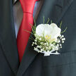 Resting just above the pocket of a dark, immaculately cut suit, this boutonnière feels like a refined nod to tradition, perfectly suited to a Mill Hill wedding or special celebration. The star of the piece is a single white orchid, its petals full and smooth, opening outwards with gentle curves that suggest poise and calm. The creamy tone of the flower glows subtly under soft lighting, while a flicker of golden warmth at its centre creates a sense of hidden depth, like a private promise shared between partners. Surrounding the orchid, a fine halo of baby's breath forms a light, cloud-like border, each tiny blossom contributing to a veil of white that enhances the bloom's purity and charm. Fresh green foliage threads through the arrangement in slender, graceful lines, reminiscent of the leafy avenues and gardens dotted around Mill Hill's residential streets. A slim white ribbon wraps the stems in a crisp, satiny band, its careful fold mirroring the structured seams of the suit's lapel. The composition is framed by a vivid red tie that cuts boldly through the image, making the white and green tones appear even more radiant by contrast. It is easy to imagine this boutonnière being adjusted with a steady hand just before walking into a ceremony at a local chapel or venue near Mill Hill Broadway, the wearer drawing quiet confidence from its understated beauty. Every element speaks of craftsmanship and attention to detail, celebrating love through subtle elegance rather than excess.
