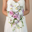 In this softly focused bridal image, the camera lingers at waist level, capturing a bride in a clean white strapless dress whose figure melts into the background so the bouquet can speak for itself. The floral arrangement is a generous, cascading design, composed in shades of white, blush pink, and muted lavender that feel at home in the gentle, village-like atmosphere of Mill Hill. Long, arching stems of white Phalaenopsis orchids pour downward from the bride's hands, their large, silky petals cupping delicate yellow-orange centres that glow subtly against the neutral beige backdrop. Gathered more closely to the bodice, plump roses in an array of pinks and soft mauves form a plush crown of colour, their petals overlapping in intricate spirals that suggest both luxury and tenderness. Tucked discreetly between these larger blooms, smaller white flowers with soft lilac edges, akin to lisianthus, play a supporting role, filling the spaces and adding a hint of ruffled charm. Fine sprays of white baby's breath weave through the bouquet like a gentle fog, ensuring that no part of the design feels heavy or overdone. Spires of pale lavender stock rise just slightly above some of the other flowers, lending slender vertical accents and a suggestion of cool, soothing fragrance-as if caught from gardens near Mill Hill Park after a light morning rain. The bouquet tapers naturally into a refined teardrop shape, guided by the orchids' downward reach and the roses' central fullness. Created with the care you'd expect from a dedicated local florist, the arrangement reflects a balanced blend of classic bridal elegance and contemporary styling, perfect for a ceremony in a local chapel or a modern venue overlooking the green hills.