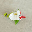 A refined boutonnière lies across a pale, textured fabric that looks like natural linen or burlap, its soft beige tone creating a calm, understated stage for the flowers. At the centre, a large white orchid bloom opens with serene elegance, each petal broad, smooth, and slightly luminous, curling gently to frame a throat touched with warm yellow and tiny orange flecks. Surrounding the orchid, fine sprays of white baby's breath are arranged in light, airy clusters, their tiny blossoms softening the silhouette and adding a sense of romance and delicacy. To the left, two striking green buds with a slightly spiky, mum-like texture stand out in vivid contrast, their fresh, energetic hue bringing modern interest to the otherwise classic palette. A single deep green leaf, likely Ruscus, extends from beneath the orchid, its glossy surface adding depth and anchoring the whole design with natural richness. The stem has been expertly wrapped with bright red satin ribbon, its sheen catching the light; the ribbon is tied into a neat little bow that rests to the right, suggesting festivity and a touch of playful charm. The overall composition feels balanced and intentional, as if prepared for a groom heading to a ceremony in Mill Hill village or a prom at a local venue near Mill Hill Broadway. You can almost sense the cool, velvety orchid against a lapel and the smooth slide of ribbon between the fingers, a small but meticulously crafted floral detail that carries the quiet excitement of an important day and the attentive work of an experienced local florist.