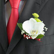 This image shows an elegant groom's boutonnière positioned on the lapel of a dark, finely cut suit, the kind you might see at a smart wedding in Mill Hill Village or a celebration close to Mill Hill Broadway. The design is anchored by a single white orchid, its petals unfurling with a serene, sculptural grace. Each petal is broad, smooth, and softly luminous, with just a glimmer of pale yellow at the centre adding warmth and depth. Behind the orchid sit two vibrant lime-green button chrysanthemums, their compact, rounded forms made of tightly packed petals that offer a lively, textured contrast to the orchid's cool simplicity. Wisps of baby's breath surround the main blooms, their slender stems branching into clusters of tiny white flowers that appear almost cloud-like, giving the piece a delicate, airy quality. Dark green leaves cradle the composition, their polished surfaces adding structure and gently framing the flowers without stealing attention. The stems are neatly bound in a slim ribbon of deep red, the satin catching the light with a soft sheen that echoes the rich red tie worn beneath the jacket. This interplay of colour-crisp white, fresh green, deep black, and warm red-creates a balanced palette that feels both timeless and contemporary. The lighting in the scene is soft, enhancing the textures: the velvet-like orchid petals, the subtle ridges of the chrysanthemums, and the feathery nature of the baby's breath. It is easy to imagine this boutonnière being prepared by a local Mill Hill florist, then carried carefully across the High Street to a waiting groom, the flowers still cool and fresh to the touch as they are pinned in place just before the ceremony begins.
