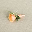 A single peach rose boutonniere lies horizontally on a softly textured light beige linen cloth, the kind of natural fabric you might see in a Mill Hill boutique along The Broadway. The rose is partially open, its petals spiralling gently from a warm, almost sunset-orange centre to creamy, shell-like edges, giving the impression of a bloom cut at the height of freshness. Nestled against its upper curve are tiny white clouds of baby's breath, each gypsophila floret no bigger than a pinhead, adding a delicate, bridal lightness around the richer tones of the rose. Slender, grass-like blades of vivid green foliage and a couple of broader, darker green leaves peek from behind the flower head, framing it with just enough contrast to feel fresh and modern while still classically romantic. The stems are neatly bound with a smooth, satin-finish ribbon in a perfectly matched peach shade, wrapped tightly and finished in a small, tidy bow that sits close to the base, suggesting careful, practiced hands at work. Light falls evenly across the arrangement, revealing the faint weave of the linen and the soft sheen of the ribbon without casting harsh shadows, creating a calm, contemplative mood. It feels like a buttonhole chosen for a Mill Hill wedding at a local church or registry office, ready to be pinned to a groom's lapel or worn by a proud parent, carrying a quiet promise of celebration and affection. The composition is intimate and uncluttered, ideal for someone who prefers subtle elegance over showiness, yet still wants their flowers to feel thoughtfully designed and personally tailored.