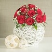 A romantic floral centerpiece, perfect for a wedding table in Mill Hill, is captured in this serene composition. At the heart of the image is a luxuriously full dome of deep red roses, between a dozen and fifteen blooms nestled closely together so their spiralled petals almost overlap, forming a plush, velvety mound of colour. Each rose is in perfect bloom, with layered petals unfurling in soft curves that invite the eye to linger. Interspersed among the roses are delicate sprays of white baby's breath, their tiny blossoms clustered like little clouds that float between the deeper tones, softening the arrangement and bringing a gentle brightness. Subtle touches of green foliage peek from within the bouquet, adding a hint of garden freshness, as if the flowers had just been gathered from a sheltered corner of Mill Hill Park. The bouquet is presented in a distinctive, round white vase built from interwoven strands that twist and overlap to create an open lattice structure, reminiscent of white wicker or rattan shaped into a sculptural orb. Its crisp white finish reflects the soft, even lighting, tying together the pure whites of the baby's breath and the decorative elements nearby. To the left of the vase, resting on the same pale, smooth tabletop, are two light, woven spheres in cream and beige tones; the larger sits slightly forward, the smaller tucked just behind, their airy construction echoing the vase's texture. Behind the arrangement, a neutral, softly textured wall fades gently into the background, lending calm and focus to the scene. The overall impression is one of quiet elegance and heartfelt romance, ideal for a wedding reception, anniversary dinner, or special occasion delivery arranged locally by Flowers Mill Hill for homes and venues across the area.