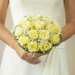 A bride clasping this delicate bouquet will feel wrapped in a soft, sunny calm: pale yellow roses, the colour of new butter warmed by morning light, form a neat, domed cluster whose velvety petals unfurl in harmonious spirals. Interwoven throughout, clouds of white baby's breath provide featherweight contrast, their tiny blossoms resting like morning dew upon the roses and making the whole composition seem lighter than its form. Around the base, artful loops of deep green grass trace an elegant outline, lending a sculptural edge that keeps the arrangement poised and compact in the hand. The scent is subtle and evocative - a faintly sweet, almost honeyed rose note tempered by a fresh, leafy green accord - as if someone had gathered a quiet patch of garden and wrapped it for the ceremony. Against a classic lace wedding dress the bouquet reads as intimate and assured, perfectly scaled for a bride walking down a chapel aisle or posing on The Ridgeway steps for photographs. As a local Mill Hill florist, I imagine delivering this arrangement for a Mill Hill Village celebration or to a family home by Mill Hill Park; the design is thoughtfully balanced to complement traditional venues and modern, pared-back styling alike. The overall mood is one of gentle joy and hopeful restraint: a floral whisper of devotion and promise that honours the ceremony without overshadowing the moment.