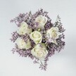 An overhead, high-angle photograph reveals a compact, hand-tied bouquet arranged with the quiet precision of an experienced Mill Hill florist. At the centre sit six creamy white roses, their petals layered in perfect spirals, tightly furled at the heart and softening towards the outer edges. Each bloom shows a velvety texture, with faint hints of pale green gathering at the base of the sepals, adding a natural freshness to the otherwise ivory palette. Around these roses drifts a halo of tiny lavender and muted lilac flowers, likely statice or limonium, forming a delicate, cloud-like ring that fills every gap without ever overwhelming the central blooms. The tiny petals and branching stems create a fine, feathery mesh, giving the bouquet a sense of airiness and depth, like a soft mist around a moon. The overall shape is circular and full, perfectly symmetrical when viewed from above, suggesting a carefully balanced design that would look at home on a Mill Hill dining table overlooking Dollis Brook or in a calm sitting room near Mill Hill Broadway. The arrangement rests on a smooth, pale surface, perhaps an off-white tabletop or studio backdrop, whose quiet, neutral tone helps the white and lilac shades stand out. Soft, even lighting falls from above, highlighting every curl of rose petal and every tiny filler flower without harsh shadows, and lending the entire scene a serene, contemplative mood. The bouquet feels suited to moments of reflection or gentle celebration-sympathy flowers sent across Mill Hill, a thoughtful anniversary surprise, or an elegant centrepiece for an intimate gathering-carrying with it a sense of purity, peace, and understated grace.