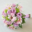 This rounded, exquisitely balanced bridal bouquet rests against a clean, neutral-toned background, its design reflecting both modern flair and timeless beauty. The various shades of pink and lavender roses unfurl elegantly, accompanied by bright, lime-green button mums that lend texture and a playful burst of colour. Trumpet-shaped pink Alstroemeria lilies with golden speckles arch gently between the focal blooms, while cloud-like clusters of delicate white baby's breath float throughout, softening the palette and exuding pure romance. Deep green leaves add freshness and natural vibrancy, peeking out to create a lush effect reminiscent of Mill Hill's verdant corners. All stems are tightly bound with a shimmery fuchsia satin ribbon, accented by a string of pearly beads-a perfect detail for weddings celebrated at local venues or intimate garden gatherings. Created by experienced Mill Hill florists, this bouquet exemplifies thoughtful artistry in every petal, making it ideal for romantic gestures, milestone anniversaries, or as an inspiration for trendsetting bridal arrangements in North London's charming Mill Hill.