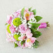 Resting on a pale, textured surface that recalls linen or fine burlap, this bouquet feels like it has been thoughtfully prepared in a Mill Hill flower studio on a calm wedding morning. The arrangement is photographed from above at a slight angle, allowing you to sense its rounded, carefully shaped form. Soft pink roses take centre stage, each flower formed from layers of gently spiralling petals that evoke timeless romance and softness. Between these roses, bright lime-green button chrysanthemums sit like little spheres of energy, their tight, almost fuzzy texture and vivid colour adding a playful, contemporary lift. Pink alstroemeria, often known as Peruvian lilies, stretch outwards with slim petals decorated by delicate markings of darker pink and soft yellow near their centres, bringing an artistic, almost watercolour effect to the bouquet. Fine sprays of white baby's breath are threaded throughout, lightening the composition and giving it a dreamy, cloud-like quality often associated with wedding posies carried down the aisle in Mill Hill churches. Around the edges, varied greenery creates a natural frame; the leaves appear smooth and lush, curving outward to give the piece volume and a fresh, garden-inspired character. The stems are brought together and wrapped securely with a luscious magenta satin ribbon, its sheen echoing the rosy tones above and adding a refined, celebratory finish. Gentle, diffused lighting enhances every detail, from the soft grain of the background fabric to the intricate veining on petals and leaves. The result is a bouquet that feels both classic and current, ideal for weddings, engagement celebrations, or same-day delivery across Mill Hill and nearby areas.