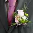 In this detailed image, a wedding boutonnière is shown with all the nuances of a piece crafted by a skilled Mill Hill florist for a special celebration. The central feature is a blush-pink rose, half-open and tender, its petals spiralling gently inwards like a soft focus point amid the more defined greenery. The rose is cradled by a selection of fresh foliage in varied shades of green, from deeper, polished leaves that give the design body, to a slim, elongated blade of foliage that traces a modern, linear shape along one side. Tiny white gypsophila flowers are scattered among these green textures, their many little blossoms forming a frothy, cloud-like veil that plays beautifully against the rose's smooth curves. Nestled at the lower part of the arrangement, a small green button chrysanthemum stands out, its neatly packed petals offering a crisp, almost graphic detail that feels current and stylish. The stems are drawn together and neatly wrapped with a soft lavender-mauve ribbon, the fabric catching the light just enough to signal celebration without overpowering the floral elements. The palette of blush, white, and fresh green would contrast elegantly against a dark or navy suit, making the boutonnière a refined choice for a groom or guest attending a ceremony near Mill Hill Broadway, Mill Hill Village, or one of the nearby chapels. The overall impression is of calm, thoughtful beauty: you can picture this being delivered early on a clear morning, ready to be pinned on as final preparations are made and the day's emotions quietly begin to build.