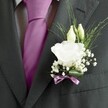 A detailed close-up of a groom's charcoal grey suit in Mill Hill, focusing on the left lapel where a refined white lisianthus buttonhole rests against the dark wool. The suit jacket has a subtle textured weave and a clean, sharply pressed lapel that curves neatly toward the camera, while beneath it a crisp white and grey patterned shirt peeks out in soft contrast. At the centre of the image, a deep magenta silk tie catches the light with a smooth, almost liquid sheen, its knot perfectly balanced between the lapels and echoing the tones of the floral ribbon below. The boutonnière itself is composed of a fully open white lisianthus bloom, its many ruffled petals layered like fine tissue, cradled by two slender ivory buds that lean gently upwards as if about to unfurl. Wisps of baby's breath scatter around the central flower, tiny white stars on filigree stems that soften the strong lines of the suit. Fine, linear greenery and a single broad green leaf curve gracefully around the base, giving structure and a fresh garden feel. Everything is tied together with a narrow mauve-to-fuchsia ribbon that mirrors the tie, wrapped snugly around the stems and finished with a discreet bow. The lighting is soft and natural, reminiscent of an afternoon ceremony at Mill Hill Park, giving the flowers a dewy, freshly arranged appearance. As a florist working locally, I'd describe this as a quietly romantic, impeccably tailored buttonhole, perfect for a groom walking up the steps of a Mill Hill church or registry office, carrying with him a sense of calm anticipation and polished elegance.