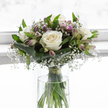 This image shows a serene hand-tied bouquet of fresh flowers, resting in a simple, cylindrical glass vase, the scene gently illuminated as if by daylight filtering through a quiet Mill Hill window. The bouquet is built around several open white roses, each one full and rounded, with velvety petals unfurling in symmetrical spirals that reveal creamy ivory tones near the centre and pure white edges at the tips. Around the roses, airy gypsophila branches create a soft veil; the baby's breath forms a mist of tiny white blossoms that drift between the larger blooms and lend an almost weightless quality to the arrangement. Nestled within this cloud are alstroemeria stems, some showing pointed buds and others partly opened to reveal slender petals brushed with lavender and pale purple hues, delicately streaked with fine lines that add a subtle decorative touch. Encasing the base of the bouquet, broad, dark green leaves curve and wrap in a structured collar, their smooth, glossy surfaces offering contrast and framing the flowers above. Finer, brighter green leaves weave upward through the centre, suggesting life, renewal, and a touch of the natural greenery found around local Mill Hill gardens and green spaces. Through the clear glass of the vase, the straight green stems are visible, neatly gathered and tied with an understated green band just above the water, reinforcing the careful craftsmanship behind the design. The background is bright and softly out of focus, with no harsh shadows, giving the bouquet a floating, contemplative presence. Overall, this arrangement feels like a quiet, respectful gesture of condolence, well suited for funerals, memorial services, or private moments of remembrance in homes and chapels throughout Mill Hill and the surrounding area.