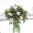 In this photograph, a tall, softly lit bouquet stands in a clear glass cylinder vase, the flowers seeming to float above the tabletop in a wash of white and soft purple tones. At its centre are creamy white roses, their petals unfolding in tender spirals that speak of purity, remembrance, and quiet love. Interspersed among them are stems of alstroemeria in gentle shades of lilac, lavender, and cool purple, some fully open, others still in bud, their slender petals edged with faint touches of green that echo the fresh foliage around them. Fine, airy sprays of white baby's breath are woven throughout, with dozens of tiny blossoms creating a delicate, cloud-like veil that softens the whole arrangement. The bouquet is framed by rich, polished greenery: broad, dark leaves provide structure and contrast, while slimmer stems add lightness and vertical lines, much like the varied greenery you might see on a peaceful walk around Mill Hill's leafy streets and open spaces. The clear water in the glass vase reveals the straight, green stems and underscores the sense of freshness and care that has gone into the design. Behind the arrangement, the background appears as a bright, softly blurred white, suggesting a light-filled room in Mill Hill where the bouquet has been placed to offer comfort. The overall impression is one of calm, elegance, and sincere condolence, making this bouquet particularly well-suited for sympathy deliveries around Mill Hill Broadway, to local churches, or to family homes seeking a gentle, floral expression of support during a difficult time.