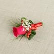 A richly detailed groom's boutonniere lies on a softly textured, light beige linen background, its natural fibres catching the gentle daylight like the tablecloths you might see in a Mill Hill garden marquee. At the heart of this small floral piece is a deep fuchsia-pink rose, its velvety petals tightly cupped at the centre and gradually unfurling, suggesting the moment just before a wedding ceremony begins. Around the rose, tiny white gypsophila blooms, or baby's breath, form a delicate halo, their airy clusters softening the bold colour and giving a cloud-like lightness to the design. Glossy red hypericum berries nestle among the petals, each bead-like berry offering a festive flash of scarlet that plays beautifully against the pink and white. Surrounding the focal flowers are layers of greenery: fine, feathery fronds that spill outward in wisps, alongside broader, darker green leaves that have been carefully curled and shaped to cradle the rose and add depth. The stems are neatly bound together with a bright red satin ribbon, wrapped smoothly and tied in a compact bow or knot that sits comfortably against the fabric of a lapel. Viewed from a slightly overhead angle, the boutonniere feels both intimate and polished, the kind of handcrafted detail a Mill Hill couple might choose for a ceremony near Mill Hill Park or along Flower Lane. The composition balances romance and subtle celebration, making it ideal as a groom's buttonhole, a groomsman's accent, or a prom accessory that quietly reflects joy, warmth, and thoughtful local florist craftsmanship.