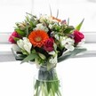 This vibrant bouquet, arranged with care in Mill Hill, is a celebration of spring's joyful promise, gathered as if from the area's peaceful gardens near Mill Hill Park. Bold orange Gerbera daisies anchor the center, their petals fanning out to create a radiant focal point that warms the arrangement with an inviting glow. Around them, creamy white alstroemeria-with distinctive curved petals and soft stripes-stretch outward, joined by richly coloured magenta roses whose gently opening petals evoke romance and quiet luxury. Soft sprays of small white chrysanthemums tiptoe through the blooms, adding delicate texture and an airy feel, while creamy and pale yellow roses bring subtle serenity. Deeper purple blossoms provide contrast, hinting at the local wildflowers that grace the area's hedgerows. All sit cradled in an abundance of glossy, broad green leaves, which sweep out to frame the bouquet and enhance its sense of fullness and movement. The arrangement gleams under soft daylight, its hues illuminated through the clear glass vase and shimmering water. Perfect for life's special moments-be it a thoughtful congratulations, a birthday, or a cheerful pick-me-up delivered to a loved one in Mill Hill-this bouquet overflows with freshness, warmth, and the sincere care of local hands.