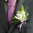 This image centres on a single, beautifully crafted boutonniere pinned to the lapel of a dark charcoal grey suit, likely worn for a wedding or prom somewhere around Mill Hill Broadway or a nearby venue. The suit fabric looks smooth and weighty, tailored with a clean line along the lapel that guides the eye towards the floral detail. Beneath the jacket sits a white dress shirt patterned with a very fine dark check, a subtle contrast that becomes a soft backdrop for a sleek lavender necktie. The tie is tied in a classic knot, the satin surface reflecting gentle light as if from a late-afternoon ceremony at a local Mill Hill church or registry office. Anchored just above the knot line, the boutonniere itself is a study in refined balance: several white Peruvian lilies unfurl with poise, their inner petals traced with delicate reddish-brown markings and touches of yellow that draw attention to their stamens. Standing tall among them is a single green, cone-shaped bud or flower spike, offering both height and an unexpectedly modern texture in fresh lime-green. Tiny clusters of white baby's breath hover around the central blooms, softening the edges like a floral mist. Slender spikes of greenery and light, grassy fronds add airiness and movement, while broader, deep green leaves frame the composition and attach it naturally to the lapel. At the stems, a lilac satin ribbon echoes the tone of the tie, tied in a precise bow that signals professional floristry. The whole arrangement suggests freshness, optimism, and the careful thought that goes into formal flowers prepared for key life moments in Mill Hill.