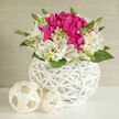 A compact and sophisticated wedding bouquet rests inside a pristine white wicker basket, radiating the kind of gentle elegance that suits a Mill Hill garden ceremony or a refined celebration near the Broadway. At the centre, a cluster of bold fuchsia blossoms forms a lively focal point, each bloom densely packed with layered petals that appear soft and almost silk-like to the touch. Surrounding this bright core is a generous halo of ivory alstroemeria, their creamy petals stretching outward in a relaxed, natural curve and detailed with tiny freckles of pale pink, adding depth and visual interest. Slender stems of greenery and a few upright buds rise between the flowers, suggesting new beginnings and subtle movement, like fresh shoots in a spring garden off Bittacy Hill. The basket itself, rounded and gleaming in pure white, is woven in a delicate lattice pattern that catches the soft light, creating shadows that feel both airy and architectural. On the smooth, pale surface beside the basket, two cream-coloured spheres made of coiled and woven material echo the texture of the handle and sides, giving the arrangement a quiet, sculptural balance. The lighting in the scene is calm and diffused, illuminating the vivid pinks and gentle whites while leaving soft, restful shadows. The overall impression is one of tranquil celebration-a design that would look perfectly at home on a wedding cake table, a guest book station, or a mantel in a Mill Hill home welcoming newlyweds back after the ceremony. The bouquet's thoughtful combination of colour, form, and texture reflects the skill of a florist who understands how to create a piece that is both striking and soothing, ideal for marking life's most heartfelt milestones.