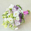 A sophisticated hand-tied bouquet rests at a gentle diagonal on a neutral light beige, slightly textured cloth, the calm background allowing every bloom to stand out clearly. The design centres on lush white lisianthus, their petals finely ruffled and almost velvety, some fully open with warm yellow hearts, others slender, tapered buds shaded in soft green and ivory, marking each stage of bloom. Between them, small clouds of pastel lavender and blush-pink flowers introduce a dreamy, airy softness, like the haze of wildflowers you might glimpse around the edges of Mill Hill's green spaces. Bold accents appear in the form of rich purple allium, each floret forming a star-shaped, spiky sphere that gives the bouquet a playful yet polished modern twist. Varied foliage provides structure and movement: elongated deep-green leaves, lighter green blades that curl and loop, and fine spiky elements that catch the light and guide the eye across the arrangement. The stems are bound tightly at the base with a magenta-fuchsia satin ribbon, smooth and glossy, creating a striking contrast with the natural greens and whites above. A fine strand of white pearls is wrapped over the ribbon, like a delicate bracelet around the handle, adding a sense of bridal luxury and careful detailing. The bouquet suggests a fragrance that is soft and refined rather than overpowering, evoking early morning air drifting over Mill Hill Broadway. Crafted with the precision of an experienced local florist, it feels perfectly suited for a wedding, civil ceremony, or elegant celebration at a nearby venue, leaving a lasting impression of calm romance, contemporary style, and bespoke elegance.