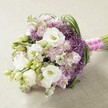 A showstopping, hand-crafted bridal bouquet from a Mill Hill florist lies elegantly atop a neutral, lightly textured setting. Billowing, ruffled white Lisianthus flowers dominate the design, their creamy petals and golden centers standing out against the delicate backdrop of unopened buds and slender, fresh green leaves. Pops of lavender and soft pink Stock are sprinkled throughout, their clustered florets offering contrast and a hint of romantic froth. Peeking through, the deep violet Allium 'Star of Persia' orbs introduce striking texture and bold visual appeal while staying in harmony with the gentle palette, much like unexpected color among the blossoms in Mill Hill Park on a summer afternoon. The bouquet's rounded silhouette is completed with a flash of fuchsia satin ribbon, elegantly wound and adorned with a fine trail of white pearls, capturing modern elegance with a timeless twist. The daylight streaming in from a Mill Hill window or garden pavilion highlights every lush detail, making this bouquet perfect for weddings, anniversaries, or milestone celebrations in NW7. This arrangement combines contemporary floristry and classic romance-an ideal choice for any occasion calling for understated luxury and fresh, local beauty.