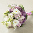 A hand-tied bouquet lies horizontally on a pale beige, finely woven fabric that looks like linen, its subtle texture softening the scene and letting the flowers shine. Dominating the arrangement are large white lisianthus blooms, their petals silky and layered, opening to reveal tender yellow-green centres that draw the eye inward, while smaller, tightly wrapped buds add a sense of promise and new beginnings. Nestled around them are abundant stems of soft pink and lilac stock, each spike packed with many little ruffled florets that bring a full, cloudlike volume and an old-fashioned, romantic charm. Scattered through the pastels are bolder accents of spiky, star-shaped purple filler flowers, similar to ornamental allium, whose intricate heads provide lively texture and a rich lavender-purple glow. Slender, glossy strands of deep green foliage, reminiscent of bear grass, thread through the bouquet and sweep in elegant arcs, especially visible along the right edge, giving the design rhythm and movement, as though echoing the gentle curves of the paths in Mill Hill Park. The gathered stems are neatly bound with a bright fuchsia satin ribbon that wraps smoothly around the handle, adding a touch of modern colour, while a single strand of tiny white pearls winds over the ribbon like a delicate bracelet. The result is a bouquet that feels airy yet luxurious, ideal for a Mill Hill bride stepping out of a local church, a registry office celebration, or a special occasion at home, where fine florist craftsmanship and soft, fragrant blooms turn a moment into a keepsake.