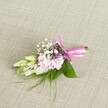 A delicate floral corsage or boutonniere rests on a softly textured oatmeal linen, the natural weave of the fabric forming a calm, neutral stage for the colours to glow. At the centre, a compact cluster of soft blush and pale pink blossoms, reminiscent of miniature stocks or small ruffled garden flowers, nestle closely together, their petals layered like gentle ripples of silk. Between the pink blooms, tiny white clouds of baby's breath add a lacy lightness, each floret crisp and bright against the pastel tones. Threaded through the cluster are subtle touches of lavender and deep purple flowers, no larger than a fingernail, which bring a quiet depth and a hint of dusk-like richness to the design. To one side, several slender, creamy white buds tinged with pale green, much like young freesias, stretch outward as if just about to open, suggesting freshness and the promise of the day ahead. Broad, glossy green leaves cradle the flowers, while finer, feathery sprays of grass-like foliage extend beyond the main shape, giving the piece a slightly wild, garden-picked charm. The stems are gathered tightly and bound in a vivid fuchsia satin ribbon, tied into a neat, symmetrical bow whose loops and trailing ends catch the light with a gentle sheen. The arrangement feels intimate and handcrafted, something you might wear at a Mill Hill wedding ceremony or prom near Mill Hill Broadway or along the leafy paths by Mill Hill Park, quietly elevating a suit or dress with a touch of thoughtful, floral elegance.