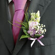 Resting against the rich, dark fabric of a tailored suit, this boutonnière appears like a soft exhale of colour and fragrance, thoughtfully composed by a florist who understands both flowers and occasion. Pale lavender blossoms, their petals brushed with cool lilac tones, gather at the centre, each one slightly cupped as though cradling the promise of the day ahead. Creamy buds peek from between the petals, offering a gentle contrast that keeps the palette light and romantic rather than bold. Surrounding this focal cluster, a collar of glossy green leaves provides structure and depth, their curved edges catching tiny highlights that suggest an indoor setting with soft, diffused light. Fine blades of greenery and delicate sprigs of white gypsophila weave through the design like notes in a quiet melody, introducing movement without disturbing the sense of calm. At the base, a sheer lavender ribbon wraps the stems, its subtle shimmer echoing the colour of the nearby tie and giving the arrangement a cohesive, well-dressed feel. The boutonnière looks perfectly at home on someone about to step into a wedding ceremony in Mill Hill, perhaps after a stroll past the shops by Mill Hill Broadway or a quiet moment in a local church porch. You can almost imagine the faint, sweet fragrance drifting upward whenever the wearer turns, a small but persistent reminder of celebration, commitment, and the care taken over even the smallest floral accessory. Crafted with the finesse expected from an experienced Mill Hill florist, this miniature arrangement becomes an intimate symbol of grace, thoughtfulness, and understated romance.