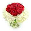 The photograph shows an elegant, dome-shaped floral arrangement placed against a clean, white background, creating a striking yet serene visual effect. At the top of the design sits a concentrated cluster of deep red roses, around nine to ten blooms gathered so closely that their petals form a continuous, rounded surface. Each rose displays the rich texture of velvety petals, some still partially closed in tight rosettes, others more open, their layered spirals unfurling gently toward the edges. This intense, romantic crown of red is gently cradled by a wide, cushion-like layer of creamy white hydrangeas. The hydrangea blooms, made up of many tiny, delicate florets, come together to form a soft, billowing base that expands outwards in a nearly perfect circle. The interplay between the bold, smooth rose petals and the fine, clustered hydrangea texture creates a sense of depth and luxury. All of the blooms are arranged in a low, clear glass bowl vase, filled with fresh water up to about halfway. Through the transparent glass, the stems are clearly visible, a neat bundle of green lines that converge toward the bottom, adding a natural, grounding element to the composition. Light glances off the glass and the surface of the water, giving the bouquet an almost luminous quality. This arrangement feels especially suited to an anniversary dinner set in a Mill Hill home, perhaps on a table near a window overlooking Arrandene Open Space or Mill Hill Park, where the calm, refined pairing of red roses and white hydrangeas can quietly underline the significance of the occasion and the tenderness of the relationship being celebrated.