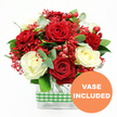 A richly romantic bouquet of red and ivory roses is photographed against a crisp white background, the style and finish echoing the thoughtful designs created for homes and businesses around Mill Hill by local florists. The red roses are the stars of the arrangement, their petals a deep, velvety crimson that opens into full, rounded blooms, each one suggesting warmth and devotion. Weaving between them, creamy white roses lend a gentle lightness, their smooth petals unfolding in soft spirals that balance the intensity of the red. Floating delicately among these focal blooms are clusters of tiny red filler flowers, reminiscent of dyed gypsophila, creating a fine, airy texture that gives the whole bouquet a sense of movement and a festive, almost frosted effect. Interspersed throughout, polished red hypericum berries add playful accents, their small, rounded shapes gleaming against the softer petals and providing an extra layer of visual interest. Around the flowers, a generous frame of greenery unfurls, with deep green foliage and subtly blue-green eucalyptus sprigs offering contrast and freshness, conjuring the peaceful, leafy feel of Mill Hill's parks and gardens. The stems converge neatly into a clear rectangular glass vase, whose transparent sides reveal the tangle of green stems and water, grounding the bouquet with clean, modern lines. Wrapped around the middle of the vase is a green and white gingham ribbon, introducing a charming country-style touch that feels welcoming and homely. In the bottom right corner of the image, an orange circular emblem carries the words VASE INCLUDED in bold white text, making it clear that this arrangement is delivered exactly as shown, ready to be placed straight on a table or countertop in Mill Hill for anniversaries, special dinners, or simply to brighten an ordinary day.