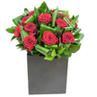Rich, velvety crimson roses form the emotive centre of this modern arrangement, each bloom opening in generous spirals that read like small, soft sculptures. The roses are carefully nested among broad, glossy green leaves that have been artfully curled and layered to create a sense of rhythm and movement; the foliage's waxy sheen catches the light and frames the crimson clusters like a living picture frame. The bouquet sits in a matte black, cube-shaped vase whose clean geometry grounds the composition and provides a chic, urban contrast to the organic forms above. Bright, even lighting sculpts the petals and highlights, allowing the velour texture of the roses to glow against the deep green of each leaf. You can almost sense the tactile contrast between the silk-smooth petal surfaces and the firmer, almost tropical feel of the foliage, and there is a subtle suggestion of fresh rose scent uplifted by green, leafy notes. Designed with intention and restraint, this piece reads as a declaration of affection or an elegant centrepiece for a contemporary dining room, office, or anniversary table. A Mill Hill florist's hand is evident in the disciplined balance between bloom and leaf, and the arrangement's compact form makes it ideal for urban spaces in and around Mill Hill Broadway, The Ridgeway or Mill Hill Village. Thoughtful and quietly dramatic, the design transforms a simple space into a memorable setting while remaining unfussy and modern.