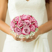 In this image, the bride holds a rounded bouquet of blush-pink roses that radiates understated luxury and calm. Each rose is fully open yet still neat, its petals curling inwards in layered spirals that create a rich, velvety texture, almost like folded silk. The pink shade is soft and romantic, a hue that would sit beautifully against the leafy avenues around Mill Hill Broadway, with slight variations from pale blush to a deeper, warm pink at the heart of each bloom. Threaded throughout the roses are airy clusters of white gypsophila, whose tiny flower heads create a gentle haze around the bouquet, softening its edges and lending an ethereal, cloud-like quality. Resting against an ivory lace wedding dress, the bouquet becomes the focal point, framed by delicate lace patterns that mirror the fine details of the flowers. The light is natural and diffused, reminiscent of a calm morning before a ceremony at a local Mill Hill church, highlighting the rose petals' subtle sheen and casting faint shadows across the bride's hands. Even without fragrance in the photograph, you can almost sense the fresh, slightly sweet perfume of the roses mingling with crisp greenery, carrying that familiar wedding-day freshness. The stems are carefully bound beneath the blooms, hidden from view, allowing the flowers to appear as a seamless dome of colour and softness. This bouquet feels timeless and composed, ideal for classic Mill Hill weddings and elegant receptions in nearby garden venues. It reflects a florist's careful eye for proportion and balance, creating an arrangement that speaks quietly of devotion, tenderness, and enduring romance.