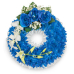 A solemn, circular funeral wreath photographed against a clean white backdrop, its main body a dense, plush ring of intensely vivid royal-blue chrysanthemums or similarly small-petalled blooms. Each electric-blue head is closely packed so the surface reads almost like velvet, catching light on countless tiny petals to create a luminous, unified sweep of colour. Around the inner edge a neat, ruffled white satin or silk ribbon frames the open centre with a crisp, formal finish, its soft sheen contrasting the textured blue. At the crown three sumptuous deep blue roses form the principal focal point; their velvety petals are fully unfurled and gently offset by feathery clusters of pure white baby's breath (gypsophila) and a few dark green leaves that punctuate the top with quiet contrast. From the left side of the wreath a graceful spray of pristine white orchids - Phalaenopsis or Dendrobium - cascades down, the orchids' broad, delicate petals and arching stems lending a refined softness that breaks the circle with an elegant movement. Intertwined with the orchids is a slender curving stem dotted with pale yellow-green buds, an emblem of new life or unfolding memory. The overall impression is dignified and consoling: a carefully balanced palette of blue and white that speaks of serenity, fidelity, and remembrance. A faint, clean floral scent seems implied in the composition, and the craftsmanship suggests an arrangement suitable for funeral chapels and memorial services in Mill Hill and neighbouring Hendon. The image conveys respectful restraint and attentive florist's care, offering a visual tribute meant to comfort those gathered to remember a life well loved.