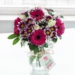A vibrant mixed bouquet sits proudly in a clear, textured glass vase, its rounded shape framed against a bright, softly blurred white background that feels like daylight streaming through a Mill Hill window. At the heart of the arrangement, bold gerbera daisies in deep fuchsia and magenta radiate outwards, each bloom with a dark central eye that draws your gaze. Around them, generously petalled chrysanthemums in purple edged with white form a rich collar of colour, their yellow centres echoing the warmth of afternoon light over Mill Hill Broadway. Nestled between these statement flowers, creamy white roses and rosebuds introduce a gentle, romantic softness, their velvety petals suggesting a light, sweet fragrance. Fine, cloud-like sprigs of white gypsophila are threaded throughout, softening the strong pinks and purples and adding a delicate, almost sparkling texture when the light catches them. Lush green foliage encircles and supports the blooms, grounding the vivid colours with natural depth. The vase itself is a squat, jar-style design, its neck wrapped with a simple bow of rustic twine, from which hangs a small white gift tag printed with a pink rose icon and flowing script, hinting at a personal message for someone special in Mill Hill or nearby Totteridge. The overall mood is cheerful and celebratory yet still elegant, the kind of fresh, professionally arranged bouquet you might have delivered to a loved one's home near Mill Hill Park for a birthday, a heartfelt thank-you, or just to brighten a kitchen table on a clear London morning.