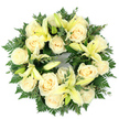 A quietly dignified circular wreath arranged flat and photographed from above, where creamy ivory roses and soft, pale yellow lilies form a tranquil ring against a clean white background. About a dozen to fifteen fully opened roses, their petals tightly swirled and satiny, create a gentle, luminous base; the roses read as classic and comforting, their subtle cream tone leaning toward warm ivory. Interspersed among them, trumpet-shaped lilies in a whisper of lemon-yellow open in graceful curves while some remain in slim, elegant buds, adding a suggestion of unfolding life and gentle movement. The flowers sit on a generous, layered bed of deep green foliage - feathery fern fronds that feather the wreath's outer edge and smaller, rounded eucalyptus-like leaves that introduce cooler, silvery-green highlights and depth. Lighting is soft and diffuse, the pale blooms catching a quiet daylight that emphasises their texture and the lilies' faint, green-tinged stamens without casting harsh shadows. The composition is deliberately dense and symmetrical, giving the wreath a full, comforting presence that reads as both formal and tender. Texturally, the contrast between the roses' velvety spirals, the lilies' smooth trumpets, and the feathery greens offers a calming interplay that can be felt as well as seen. The circular form and serene palette of cream, lemon and green make this a classic memorial design - a visual expression of peace, purity, and remembrance appropriate for a funeral service, graveside tribute or a quiet gesture of condolence for someone in Mill Hill or nearby communities seeking a respectful, floral farewell.