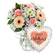Captured against a fresh white background, this Mother's Day display feels like a bright, heartfelt surprise waiting on a hallway console in Mill Hill. At the centre is a lush mixed bouquet arranged in a clear glass vase whose smooth, simple lines let the flowers take all the attention. Around the neck of the vase, a band of rustic twine is tied, adding a reassuringly homespun touch that suggests it was prepared by hand rather than by machine. Rising from the vase is a harmonious blend of pastel blooms: several peach-pink roses open wide, their velvety petals arranged in elegant spirals that lead into softly shadowed centres. Beside them, broad peach gerbera daisies with dense rings of petals and strong dark brown centres provide a cheerful, contemporary counterpoint, adding a sense of fun and optimism. Sprays of delicate white baby's breath thread their way through the arrangement, like tiny foam bubbles resting on the petals, while smaller white chrysanthemums with apple-green hearts fill the spaces, giving the bouquet a rounded, abundant feel. Rich green foliage is interlaced among the flowers, providing a cool, leafy backdrop that enhances the warmth of the peach and pink tones. To the right of the bouquet, gently leaning as though caught mid-float, a heart-shaped Mylar balloon completes the gift. Its surface is bright white and scattered with small metallic gold and silver dots that catch the light like sequins. In the middle of the balloon, a warm peach heart frames the words HAPPY Mother's DAY, printed in bold black lettering, with the word Mother's rendered in a soft, flowing script that echoes the curves of the rose petals. A small white greeting card peeps out from behind the arrangement, hinting at a personal message inside. Overall, the image conveys a warm, celebratory mood, ideal for showcasing Mother's Day flower delivery throughout Mill Hill, from homes near Mill Hill East Station to leafy streets off The Ridgeway.